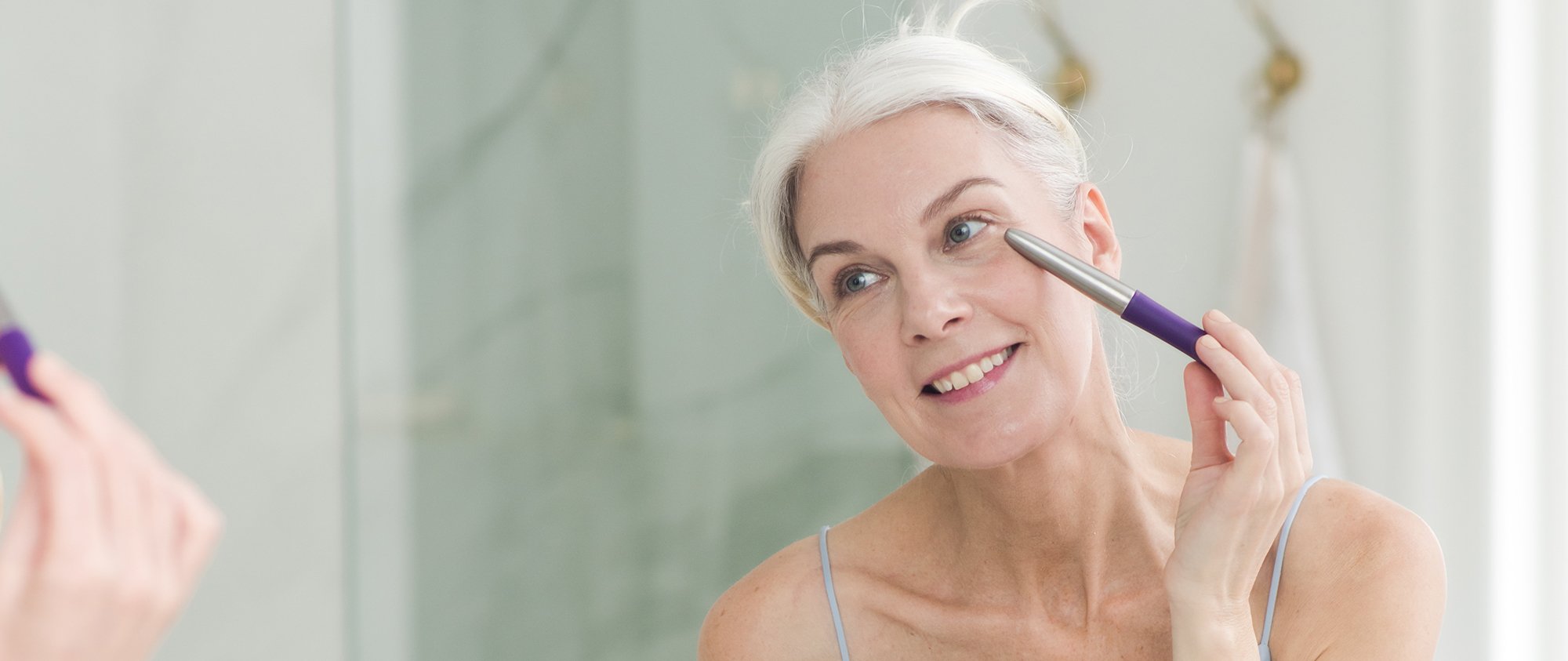 Silver-haired woman using a facial massage wand beneath her eye while looking in a mirror.