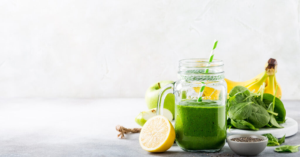 A green smoothie in a glass jar beside spinach, bananas, apples, lemon, and chia seeds.