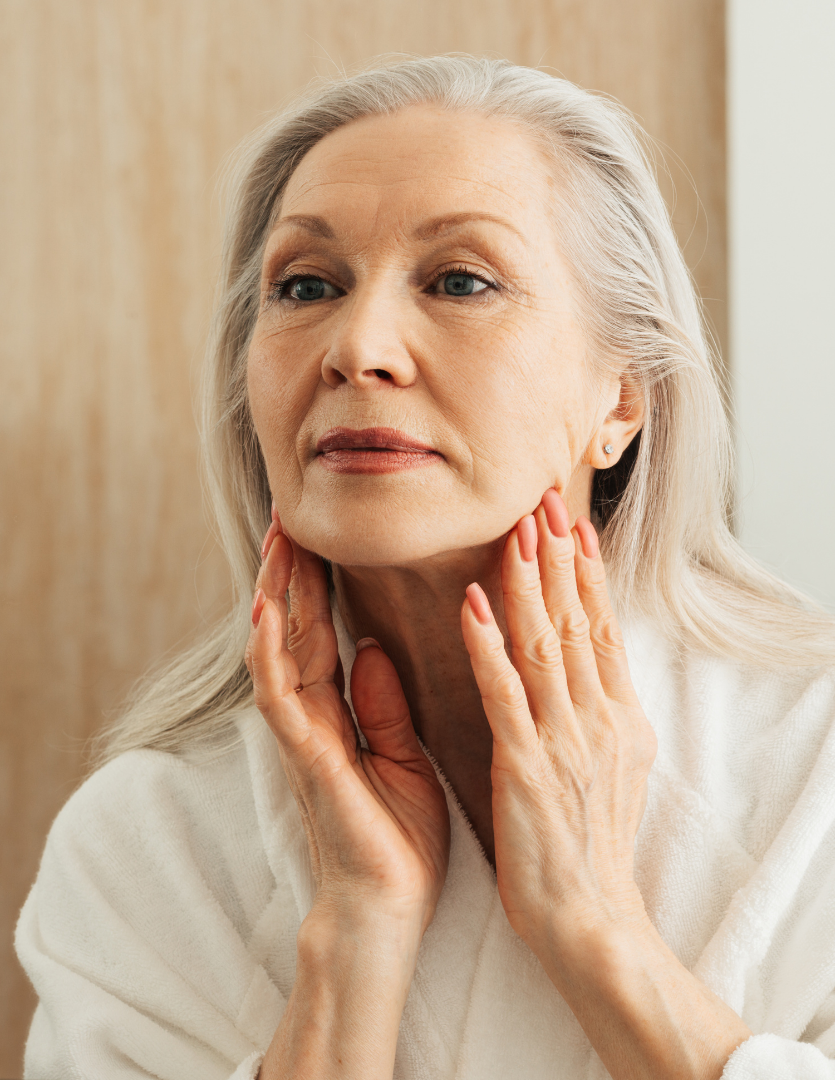 Silver-haired woman in a white robe gently touching her jaw and neck.