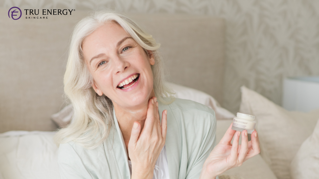 Smiling silver-haired woman holding a jar of Tru Energy cream and touching her neck.